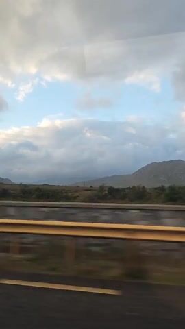 The video shows a time-lapse of a landscape from a moving vehicle. In the foreground, there is a yellow barrier and a dark road. Beyond the barrier, there is a field with green and brown vegetation. In the background, there are mountains under a cloudy sky. The clouds appear to be moving and changing shape, suggesting the passage of time. The lighting suggests it is either early morning or late afternoon.
