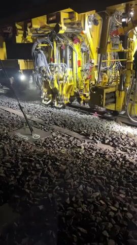 A yellow railroad maintenance machine operates on a track at night. The machine has multiple hydraulic arms and mechanical components. Lights on the machine illuminate the track and surrounding ballast. The machine appears to be vibrating and moving slowly. One of the lights on the machine is directly above the tracks. The ground is covered in ballast stones. The machine has the word "UNIMAT" visible on its side. The video is shaky and appears to be filmed handheld.