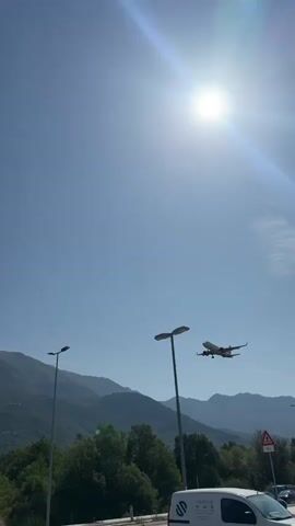 A plane with red and white livery flies across a clear blue sky, with the sun brightly shining in the upper right corner. The plane appears to be on approach for landing. Below the plane, a range of mountains covered in green trees forms the background. In the foreground, there are two lampposts and some palm trees. A white van with a logo on its side is partially visible at the bottom right of the frame.
