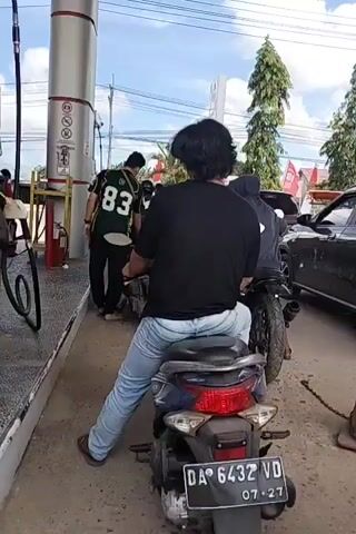 A group of people are at a gas station. A man wearing a black shirt and blue jeans is sitting on a motorcycle, facing away from the camera. Another person wearing a dark hoodie is behind him, also on a motorcycle. A dark grey car is parked next to them, and a man wearing a black and green shirt with the number 83 is standing next to it. There are traffic cones with chains on the ground. The gas station has tall white columns and red signage. Trees and a cloudy sky are visible in the background.