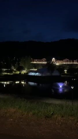 The video is a dark, low-light shot of an outdoor scene at night. The foreground is dominated by a grassy, dirt embankment. In the middle ground, there is a body of water with a fountain illuminated by a purple light. On the far bank of the water, there are several white tent-like structures. In the background, a large, multi-story building complex is visible, illuminated by numerous warm-toned lights arranged along its facade and roofline. The building sits at the base of a dark, forested hill. The sky is a deep, dark blue, with minimal visible cloud cover. The overall impression is of a quiet, illuminated landscape at night.