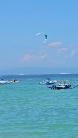 A kiteboarder is visible in the sky over a body of turquoise water under a clear blue sky. Several traditional outrigger boats are floating on the water. One boat is in the foreground with blue trim, and another is behind it. Further out in the water, a person is riding a jet ski. In the background, a distant shoreline with mountains is visible under a hazy sky.