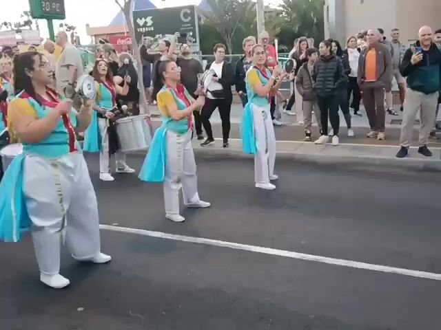 A group of about ten women are marching on a street in a parade. They are wearing matching costumes with light blue, yellow, red, and white colors. Some are playing snare drums while others are playing tambourines. Spectators line both sides of the street, watching and filming the parade with their phones. A large billboard is visible in the background, along with palm trees and buildings. The temperature is displayed as 20 degrees Celsius.