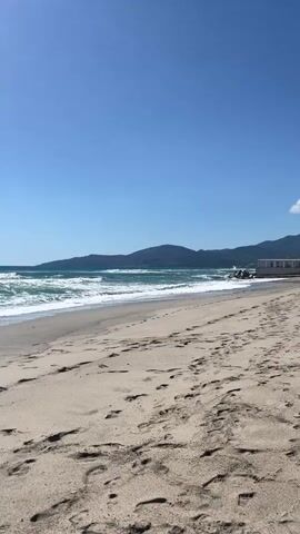 The video shows a beach with several footprints in the sand, leading toward the ocean. The water is a dark turquoise color and there are many small waves. In the background, there are some mountains. In the distance, there appears to be a person swimming. The sky is clear and blue with the sun shining brightly.