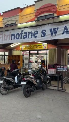 The video shows the exterior of a small store with "SELAMAT DATANG" (Welcome) written on a banner above the entrance. Several motorcycles are parked in front of the store. Inside, there are shelves stocked with various items, including water bottles and what appear to be cigarettes. Three women and a young girl are visible inside the store. One woman, wearing an orange shirt, is leaning over a counter, appearing to work. Another woman in a headscarf is looking at items on a shelf. A third woman, also in a headscarf, stands near the counter. The young girl is standing next to the woman in the orange shirt. The lighting suggests it is either dusk or nighttime.