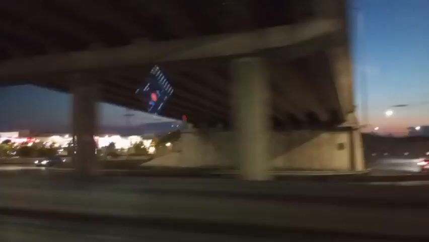This video shows a view from inside a vehicle, likely a car, moving along a road. The scene is captured at dusk or early evening, with a dark blue sky transitioning to orange and pink near the horizon, indicating sunset or sunrise. Above the road, there is an overpass or bridge structure, with concrete pillars and a concrete ceiling visible in the foreground. Through the gap under the overpass, a commercial area is visible.

Prominently, a tall, illuminated sign or tower stands out in the distance, displaying vertical yellow lights. In front of it, there are buildings with illuminated facades, suggesting a shopping center or entertainment complex. Numerous smaller lights, possibly streetlights and building lights, are spread across the scene, creating a festive or lively atmosphere. Trees and landscaping are also present in the illuminated area.

On the road below the overpass, there are other vehicles. Headlights of oncoming cars are visible as streaks of light, and some cars appear to be in motion. The road surface is dark asphalt. Reflections are visible on the windshield of the vehicle from which the video is taken, including what appear to be lights and possibly the interior of the vehicle. The overall impression is a nighttime drive past a brightly lit commercial district under a twilight sky.