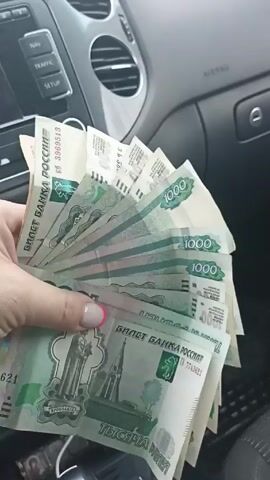 Here is a description of the video:
A hand is seen inside a car, holding several 1000 Russian Ruble banknotes. The hand, featuring a manicure with pink accents, gently fans the bills, displaying them to the camera. The background shows a car interior, including dashboard controls and air vents, suggesting the scene is taking place inside a vehicle. The lighting appears to be natural, highlighting the green color of the banknotes and the textures of the car's interior.
