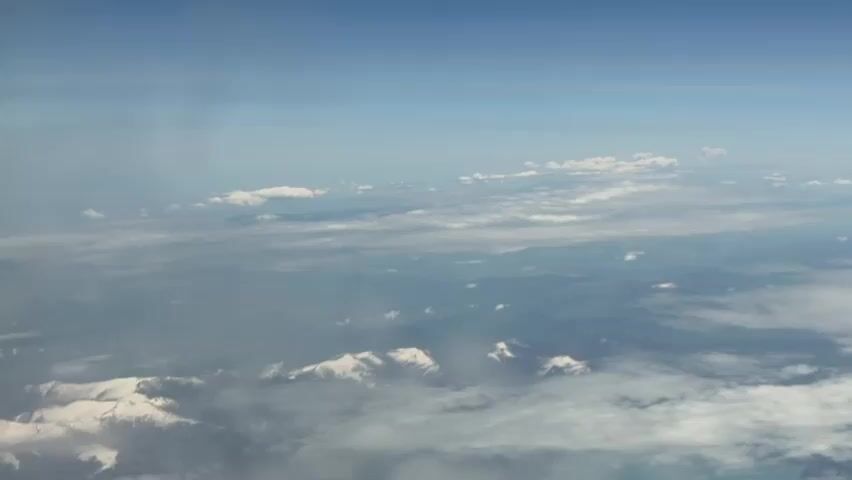 The view from an airplane window shows a vast landscape of mountains covered in snow. Above the mountains are scattered clouds against a clear, blue sky. The scene is calm and peaceful.