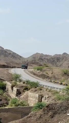 A semi-trailer truck drives along a paved road that curves through a barren, mountainous landscape. The truck is dark green with a lighter colored trailer, and it is traveling away from the camera. The road has a white line marking the center and a white guardrail with yellow reflectors along the right side. The surrounding terrain is rocky and arid, with sparse vegetation consisting of small trees and shrubs. In the foreground, there are remnants of stone walls, suggesting old structures or landscaping. The sky is clear and pale blue, indicating a daytime scene with dry, possibly hot weather. The overall impression is one of a remote, rugged environment.