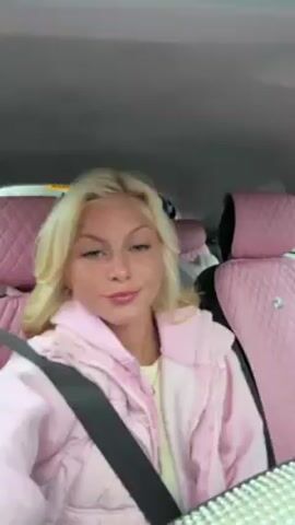 A young woman with blonde hair is in a car wearing a pink jacket. The car interior is also pink with a diamond-stitched design on the seats. She is wearing a seatbelt. She looks at the camera and mouths words. Music is playing. The woman appears to be the focus of the video. The location is inside a vehicle.