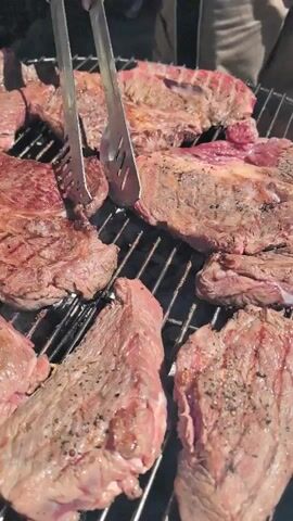 The video shows several raw steaks on a charcoal grill. A person's hands are using tongs to flip the steaks, which are seasoned with salt and pepper. The grill is outdoors, and the background is blurred. The steaks are various sizes and cuts, and they appear to be cooking evenly. The video is shot from a close-up perspective, focusing on the steaks and the grilling process.