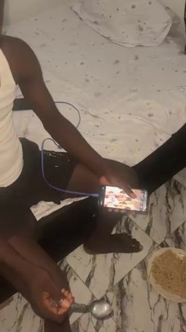 A person is sitting on a bed with a phone in their hand, which is plugged into a charger. They are wearing a white tank top and black shorts. There is a spoon in their other hand and a bowl of food on the floor. The room appears to be a bedroom with a patterned bedspread. The person looks at the phone and then at the camera.
