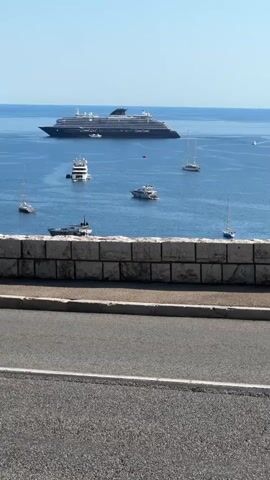 This media file captures a scenic view of Villefranche-sur-Mer, France, on a bright, sunny day. The primary subjects include a large cruise ship, several yachts and sailboats, and various vehicles moving along a road overlooking the water.

In the foreground, a stone wall and asphalt road are visible. A black convertible car drives past, with its top down, revealing two occupants. Following the car, a man and a young girl ride a scooter. The man is wearing a helmet and a t-shirt, and the girl is wearing a helmet and a dress.

The middle ground showcases the beautiful bay of Villefranche-sur-Mer, dotted with numerous boats of various sizes, including a prominent cruise ship in the distance. The water is a clear blue, and the coastline is lined with lush green hills and buildings, suggesting a Mediterranean setting.

The background reveals more of the hillside town and the expansive blue sky, indicating fair weather and a clear day, likely during daylight hours. The overall atmosphere appears relaxed and idyllic, typical of a coastal vacation spot. No visible text is present in the media file.