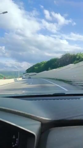 A front-facing, dashcam view shows a vehicle driving on a highway on a cloudy day. The dashboard is visible in the foreground. A few vehicles are seen in the distance on the highway. To the right of the highway, there is a tall, light-colored wall with green trees and bushes behind it. To the left, there are streetlights and distant mountains.