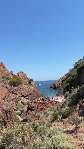 The video shows a scenic view of a rocky coastline with a clear blue sky and calm ocean water. Several people are visible on a small beach, some relaxing and others swimming or wading in the water. There are some boats visible in the distance on the ocean. The vegetation is sparse, consisting of low-lying bushes and some trees on the rocky outcrops. The rocks are reddish-brown in color, suggesting a geological formation common in certain coastal areas. The overall impression is one of a peaceful summer day at a secluded beach.