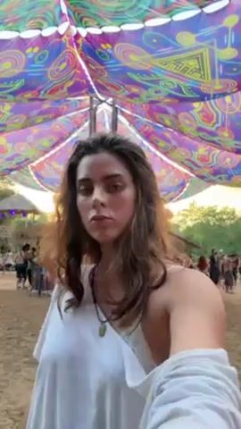 A young woman stands in a festival setting, directly facing the camera and lightly dancing. She is wearing a white t-shirt draped off one shoulder, revealing a white lace bra underneath. She has long brown hair and a necklace. She puts her hands in her hair before continuing to dance. Behind her, people mill about on a sandy ground, and there are trees and some structures visible. Above her, a colorful, patterned fabric canopy is strung up. The overall atmosphere is that of a party or outdoor gathering.