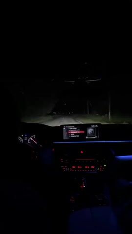 The video shows the interior of a car at night, with the dashboard illuminated. The car appears to be in motion, as suggested by the illuminated headlights in the distance. The dashboard features a digital display screen and various buttons and controls, many of which are lit in red and blue. The overall impression is one of a nighttime drive through a dark, possibly rural, area.
