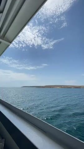 The scene is captured from a boat sailing across a body of water towards a somewhat barren, rocky coastline under a partly cloudy blue sky. The water is moderately choppy, reflecting sunlight. The camera is positioned near the edge of the boat, with parts of the boat's structure visible in the foreground. A small bird flies across the frame. The overall environment conveys a sunny day on the water.