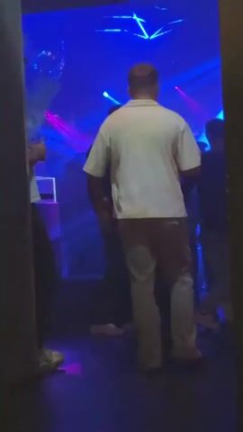 The video shows three people standing in a nightclub environment. The lighting is dynamic, shifting between purple and blue hues. One person is visible on the left, holding a drink, partially obscured by what appears to be a doorway or frame. Two other individuals, further inside the club, are seen with their backs to the camera. One is wearing a dark jacket, and the other is wearing a light-colored short-sleeved shirt and grey pants. There is a crowd of people further inside the club. The scene is dimly lit and filled with dance music.