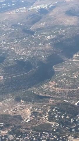 The video displays an aerial view of a rugged, hilly landscape, possibly taken from an airplane. The terrain is sparsely populated with scattered buildings, some appearing as clusters of houses and others as isolated structures. Roads wind through the hills, connecting these settlements and traversing the natural contours of the land. The vegetation is dry and scrubby, suggesting a warm, possibly arid climate. Sunlight casts shadows across the landscape, indicating a clear day and casting doubt on the time of day. The scene evokes a sense of remoteness and perhaps a touch of desolation due to the arid environment and the scattered nature of human habitation. There are no visible people or distinct objects of significant interest, beyond the settlements and infrastructure. The overall impression is one of a natural landscape shaped by human presence in a quiet, unassuming manner.