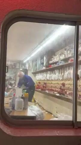 An eye-level, exterior shot through a window shows an older man working at a counter inside a store or shop. The man, with white hair and wearing a blue jacket, appears to be focused on an object on the counter. The counter is cluttered with bottles, containers, and paper towels. Behind the counter, shelves are lined with numerous items, some appearing to be white gloves or similar objects, arranged in rows. The window is somewhat dirty and the exterior wall surrounding the window is textured and painted red.  The interior of the store is brightly lit.