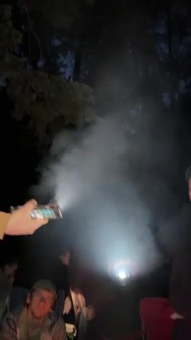 In a nighttime forest setting, several people are gathered around a campfire. The image is dimly lit, with the primary light source coming from a flashlight held by a person in the foreground, whose arm is visible on the left side of the frame. The flashlight beam cuts through the smoke from the campfire, creating a visible shaft of light. Another person is holding a flashlight towards the smoke from the right side of the frame. In the foreground, a young man wearing a camouflage jacket and a green baseball cap is seated and looking at the camera. Other individuals are partially visible in the background, seated in camping chairs. The trees in the background are silhouetted against the dark sky, suggesting it is nighttime. The atmosphere appears to be one of a casual outdoor gathering or camping trip.