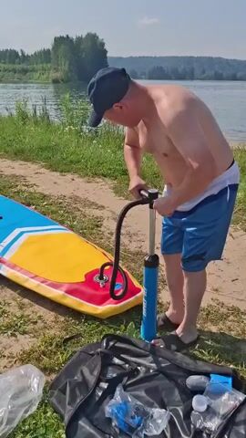 A shirtless man wearing blue shorts and a black baseball cap is pumping up a red, yellow, blue, and white inflatable stand-up paddleboard with a blue hand pump. He is standing on a sandy and grassy shore next to a body of water. A black bag and other items are on the ground near him.