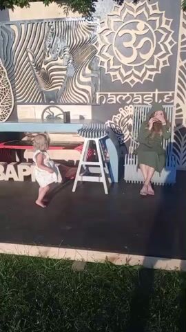 A little girl in a white dress is standing to the left of a stool with a striped top. A woman in a green hooded dress is sitting on a chair to the right of the stool. The woman is covering her face with her hands. Behind them is a wall decoration with an om symbol, the word "namaste", and a pattern of leaves. The ground is black, and there is grass in the foreground.