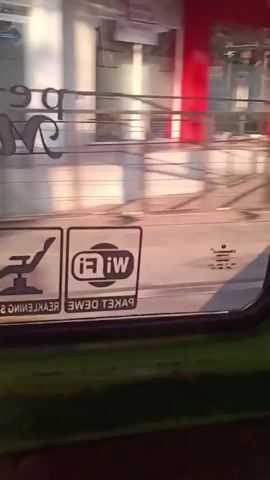 The video shows a view through a vehicle window, likely a bus or train. Outside the window, there is a blurry street scene with buildings and trees. On the window itself, there are two stickers. One sticker depicts a reclining chair with a line through it, possibly indicating no reclining seats. The other sticker shows the Wi-Fi logo and text that appears to be in a foreign language. The text on the stickers is reversed as viewed from inside the vehicle. The video appears to be recorded while the vehicle is in motion, causing the external environment to be out of focus.