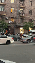Crowd gathers on city sidewalk as traffic passes