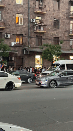 Crowd gathers on city sidewalk as traffic passes