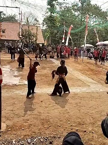 The video shows an outdoor cultural performance. In the foreground, two individuals are engaged in a stylized fight. One person, wearing a dark outfit and a headdress, holds a whip. The other person, in a maroon shirt and dark pants, wields a stick. They are on a dirt arena, surrounded by spectators. In the background, more performers are present, along with flags and what appears to be a stage with speakers. The environment is rural, with trees and buildings visible. A close-up shot at the end shows a person in a red garment with what appears to be a rooster in their hands.