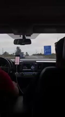 The video depicts an interior view from the back seat of a car driving on a highway. Two people are visible: the driver, wearing a red shirt, and another passenger wearing a black shirt in the front passenger seat. The dashboard is visible, featuring a digital interface with what looks like radio information displayed. A smartphone is mounted on the dashboard acting as a GPS. The highway stretches out in front of the car, with other vehicles present. Roadside vegetation can be seen alongside the road, and a road sign indicating directions to Temuco, Lautaro, and Quepe is visible on the right. The sky is overcast.