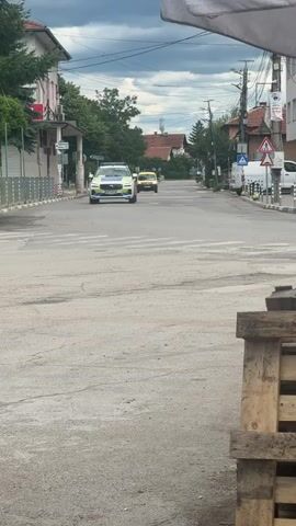A white Volvo police SUV with blue and yellow markings and lights is driving down a paved street in a town. A yellow car is visible further down the street behind the police SUV. The street is lined with buildings and trees on both sides. There are traffic signs and streetlights along the road. In the foreground, there is a wooden pallet. The sky is overcast.