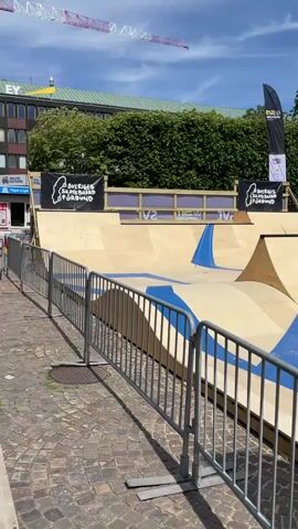 The video pans across a temporary outdoor skatepark constructed from light-colored wood with blue accents. A metal fence lines the foreground. In the background, buildings and trees are visible, along with construction cranes. A man is seen crouching near the skatepark, appearing to work on a plan or diagram. Flags and banners indicating an event are displayed. The sky is blue with some clouds.