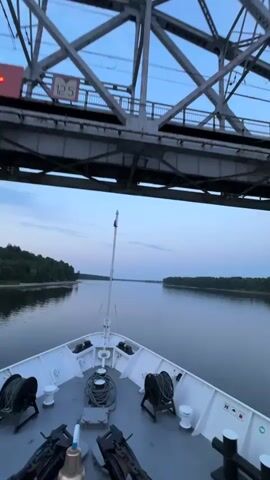 The video begins by showing the bow of a white ship as it travels under a large steel railway bridge. The sky is a deep blue, transitioning to a lighter shade near the horizon, with some clouds visible. On the bridge, a sign with the number 12.5 is visible. The camera pans up and to the right, revealing the underside of the intricate steel structure of the bridge. The water is calm and reflects the sky. As the ship moves forward, a person is seen standing on the deck of the ship, looking out at the bridge. The camera continues to pan, showing more of the bridge's structure and its support pillars in the water. The banks of the river are lined with trees, and in the distance, a few buildings are visible on the far bank. The video ends with a wide shot of the bridge spanning across the river, with the ship still moving beneath it.