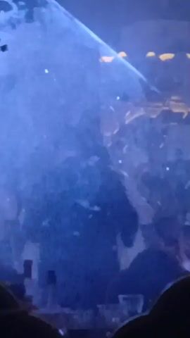 A couple, one in a white wedding dress and veil and the other in a dark suit, are kissing in a dimly lit room. A blue light, possibly a spotlight or laser, illuminates the scene from the left, casting a haze of smoke or mist. The background is indistinct but appears to be an indoor venue with some decorative elements, including what look like hanging golden fixtures. Several people are visible in the foreground and background, out of focus, suggesting the video was taken from within a crowd.