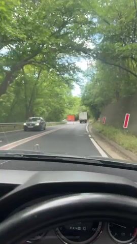 The video shows a first-person view from inside a car driving on a winding road. The road has a double yellow line in the center and is lined with trees on both sides. There are two trucks ahead, one red and one white. Orange pylons are placed in the center of the road. A speed limit sign is visible on the right side of the road. The sky is partly cloudy.