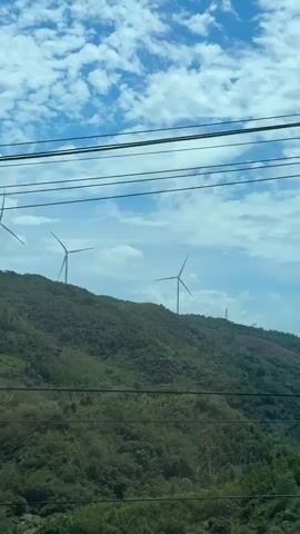 The video captures a scenic view of a lush, green mountainside dotted with wind turbines. The sky is a vibrant blue, filled with fluffy white clouds, suggesting a pleasant day. The camera appears to be moving, possibly from a vehicle, as indicated by the blurred foreground foliage and the presence of power lines stretched across the frame.

The primary subjects are the wind turbines, tall structures with rotating blades, strategically placed on the undulating terrain. Their presence signifies a renewable energy source operating within a natural, rural environment. The mountains themselves are densely covered in vegetation, with various shades of green indicating a healthy ecosystem.

The activity taking place is the natural phenomenon of wind powering the turbines, a silent but continuous process. The camera's movement provides a dynamic perspective, showcasing the expanse of the landscape and the distant turbines.

Notable details include the clear blue sky with scattered clouds, hinting at fair weather conditions. The time of day appears to be daytime, likely late morning or afternoon, given the brightness and the shadows cast on the mountainside. The location, Đakrông Commune, Vietnam, further contextualizes the tropical or subtropical climate suggested by the abundant greenery. There are no visible text elements in the video. The overall mood conveyed is one of natural beauty and sustainable energy, creating a sense of peace and environmental consciousness.
