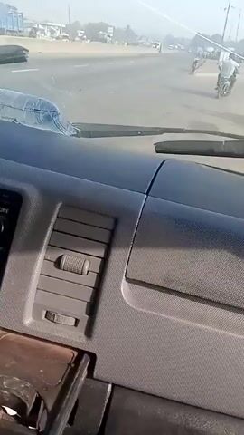 The video shows the interior of a vehicle driving along a road. The dashboard is visible, with a radio, air vents, and a cupholder. A bottle of water is placed in the cupholder. Through the windshield, the road and surrounding environment are visible, including buildings, other vehicles, people on bicycles, and street signs. The video seems to be taken from the driver's perspective. The video is shaky. The weather appears sunny.