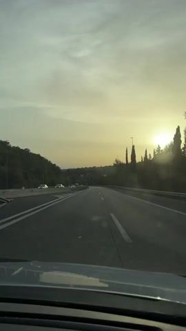 The video shows a view from inside a car driving on a highway. The sun is low in the sky, casting a golden glow and creating lens flare effects. The highway has multiple lanes, and there are other cars visible. On the left side of the road, there is a grassy embankment with trees and guardrails. On the right side, there are more trees and some tall, slender structures that might be streetlights or decorative elements. The sky is partly cloudy. The overall impression is of a scenic drive during sunset or sunrise.