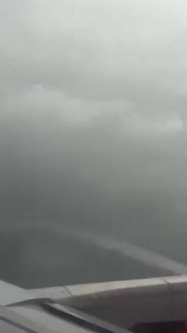 The video shows a view from the window of an airplane. The plane's wing is visible in the foreground. The sky outside is filled with thick, gray clouds, creating an overcast and gloomy atmosphere. The clouds appear dense and uniform, obscuring any view of the ground or sky beyond.
