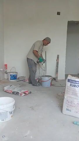 A man wearing a light brown shirt, grey pants, and a grey skullcap is standing in a room that appears to be under construction. He is holding a tool with a long shaft and a mixing attachment, which he is using to mix something in a red bucket. His hands are gloved in light green. The floor is concrete and littered with construction materials, including bags of powdered material, plastic buckets, and coiled electrical cords. In the background, there is a white wall, a doorway leading to another room, and various construction tools and supplies. The lighting suggests an indoor environment with artificial light or diffused natural light.