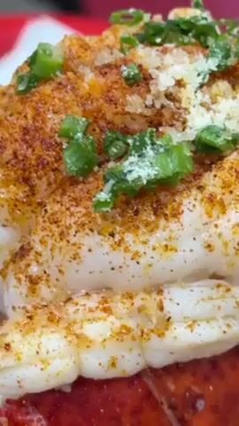 The video shows a loaded baked potato topped with lobster meat, spices, and green onions, placed on a red tray.  A person is seated at a table in what appears to be a casual dining area. The baked potato is served on a white paper plate. The topping includes a lobster tail on top, with what looks like shredded crab and a lemon wedge on the bottom half of the potato.  Salt packets are also visible on the tray. There are other tables and chairs in the background, along with posters on the wall.