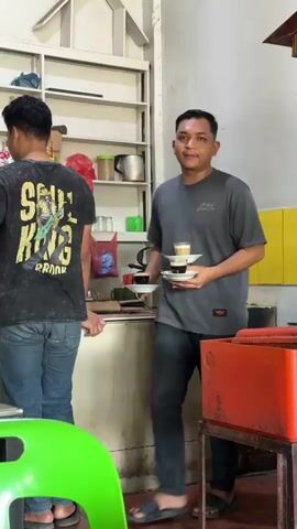 A man in a grey t-shirt and jeans is holding two small cups of coffee in his hands. He is standing in front of a shelf filled with bottles and cans. Behind him, another man in a black t-shirt and jeans is visible. The environment appears to be a shop or cafe.
