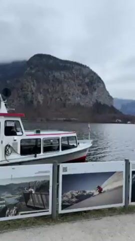 The video shows a white boat with a red stripe along its hull, docked in a lake. There are mountains in the background, partially covered with snow, under a cloudy sky.  In the foreground are two information boards with photos on them.