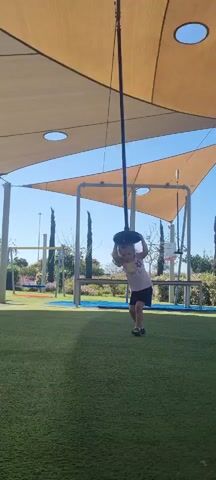 A young child is on a playground holding onto a vertical pole with a disc at the top. They are standing on green artificial turf beneath a shade structure made of large beige fabric stretched over metal frames. The sky is bright blue. The child is wearing a light-colored T-shirt and dark shorts. They are alternately reaching up and letting the disc swing down. In the background are more playground elements, trees, and a tall utility pole in the distance.