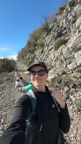 A woman with sunglasses and a cap smiles at the camera on a rocky mountain path. Another woman is behind her, taking a photo. The camera then pans to show a view of a body of water, possibly a lake or sea, surrounded by mountains. The sky is blue with scattered clouds, and the sun is shining brightly. The landscape is rugged with rocky terrain and sparse vegetation.