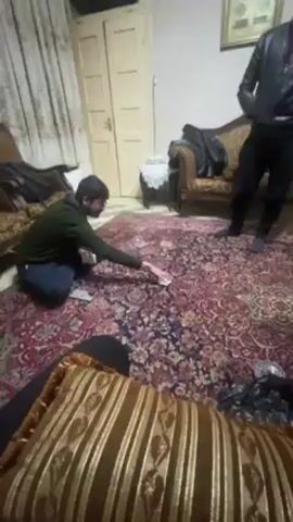 A man is seated on the floor with his legs crossed, dealing cards on an ornate rug. Another man in a leather jacket stands near the center of the rug, gesturing with his hands and speaking to the other man. A third man in a gray jacket walks around the rug, also speaking. The rug covers a large part of the room, and there are two ornate chairs, a small table with a white cloth, and a framed picture on the wall in the background. A striped pillow is in the foreground.