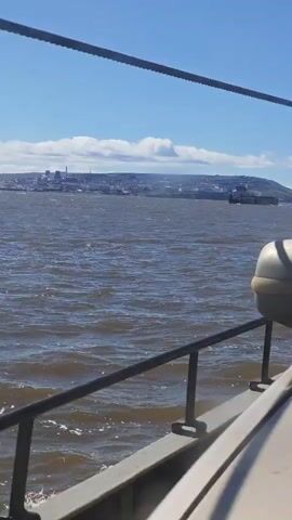 A video taken from the perspective of a boat shows a wide expanse of choppy, brown water under a bright blue sky with scattered white clouds. In the distance, a city skyline with buildings and what appears to be industrial structures is visible along the coast of a large hill. A cargo ship is seen moving in the water between the boat and the city. The foreground of the video shows the side of the boat, including a railing and part of a large, light-colored object, possibly a life raft or storage container. The boat appears to be in motion, with a slight wake visible in the lower left corner.